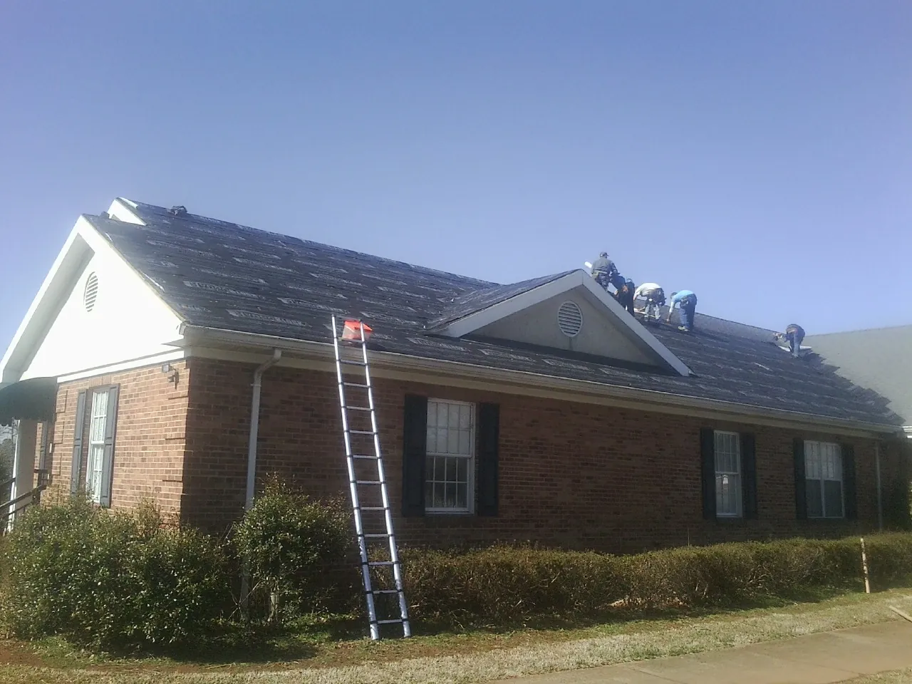 Crew tearing shingles off a brick ranch roof with a ladder set — Emergency Roof Repair work by our crew in Roopville