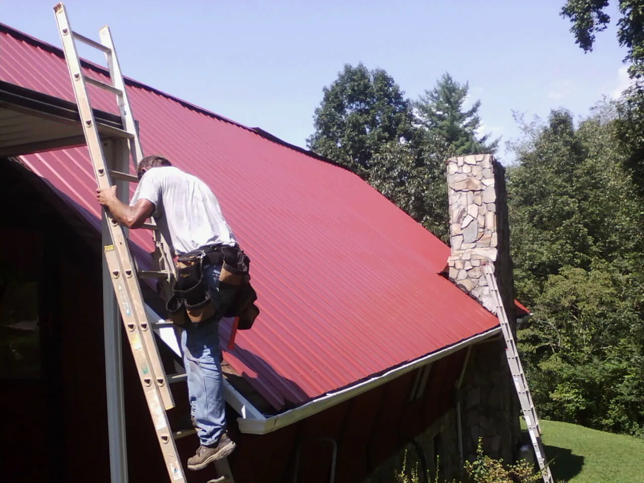Roofer on a ladder beside a red metal roof and stone chimney — Soffit & Fascia Repair by Roofing Brothers in Roopville